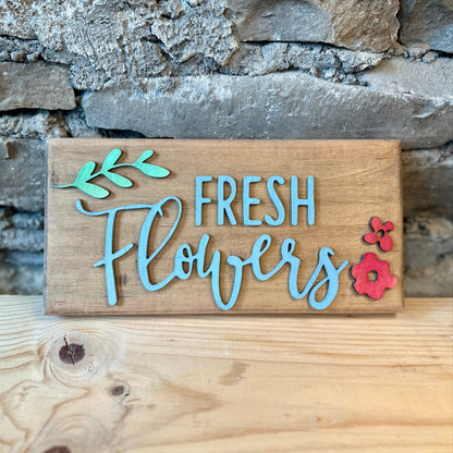 Wooden sign saying 'Fresh Flowers' with leaves and blossoms by a stone wall.