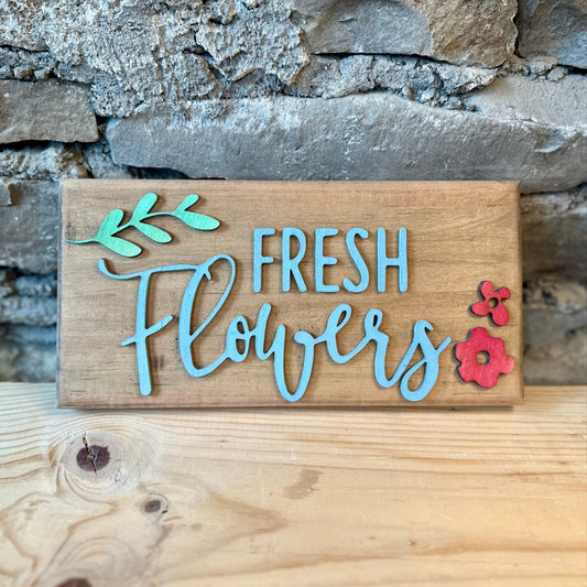 Wooden sign saying 'Fresh Flowers' with leaves and blossoms by a stone wall.