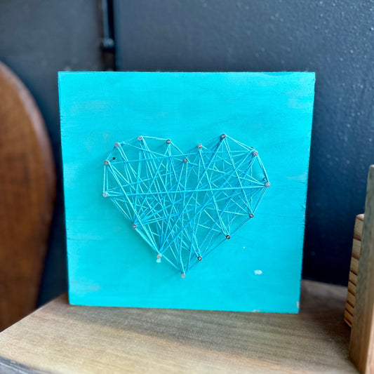 Turquoise board with a string art heart design on a wooden shelf.