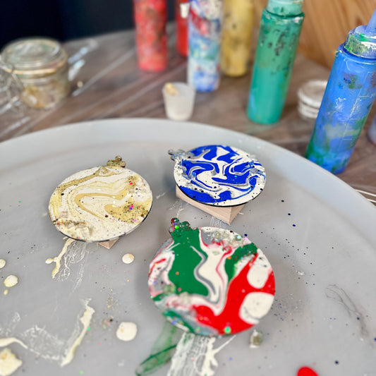 Decorative wooden ornaments with colorful designs on a tray, with paint bottles in the background