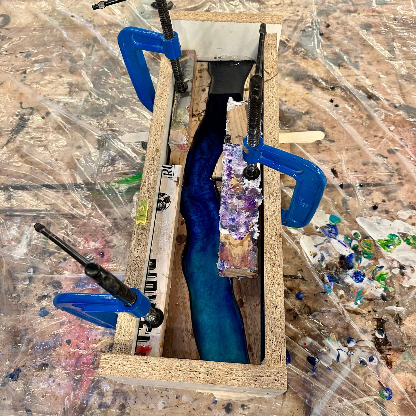 Workbench with a wooden frame holding a blue and wood epoxy river coaster set with blue clamps.