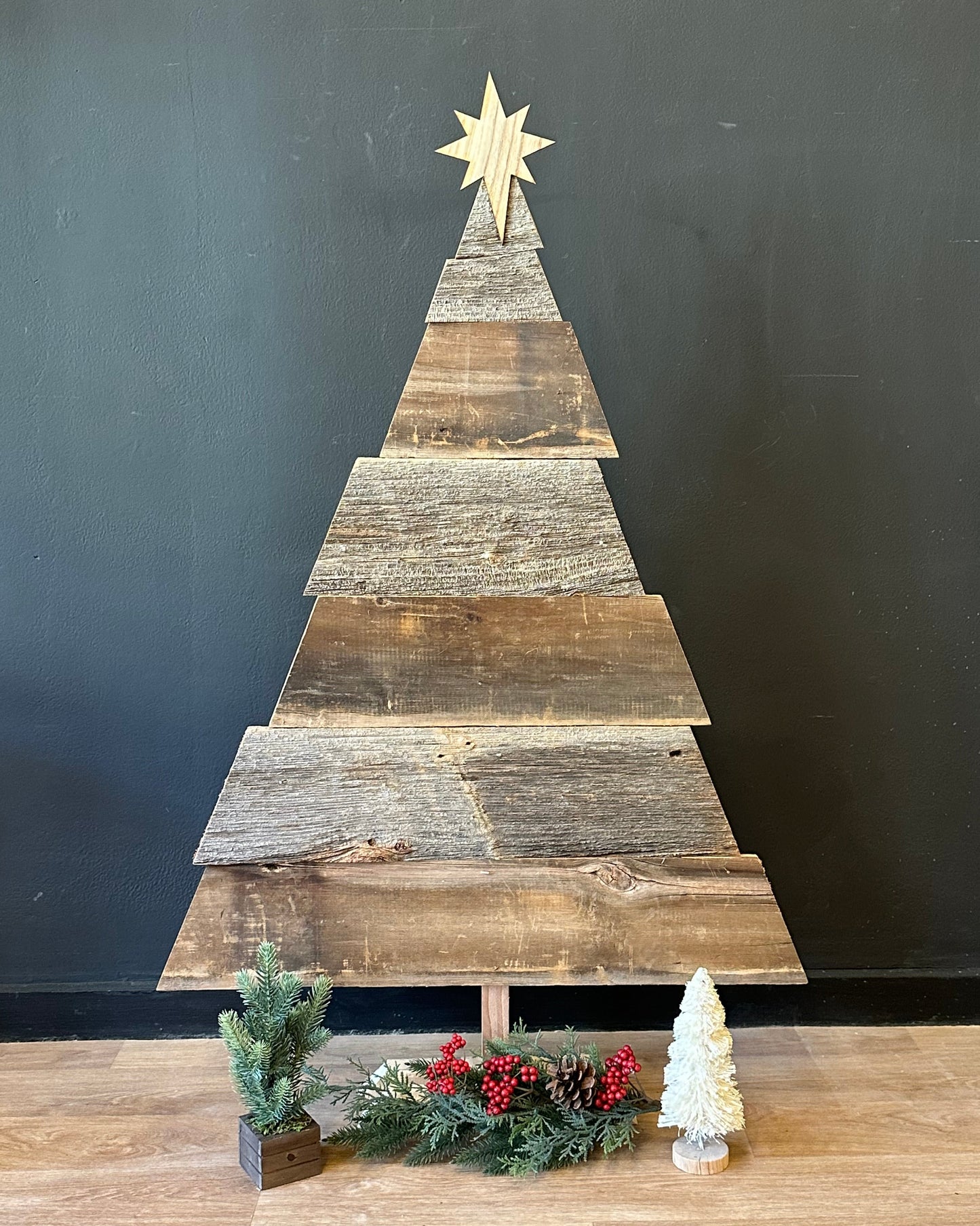 Wooden Christmas tree against a black wall with a star on top, next to a decorated Christmas tree on a wooden floor.