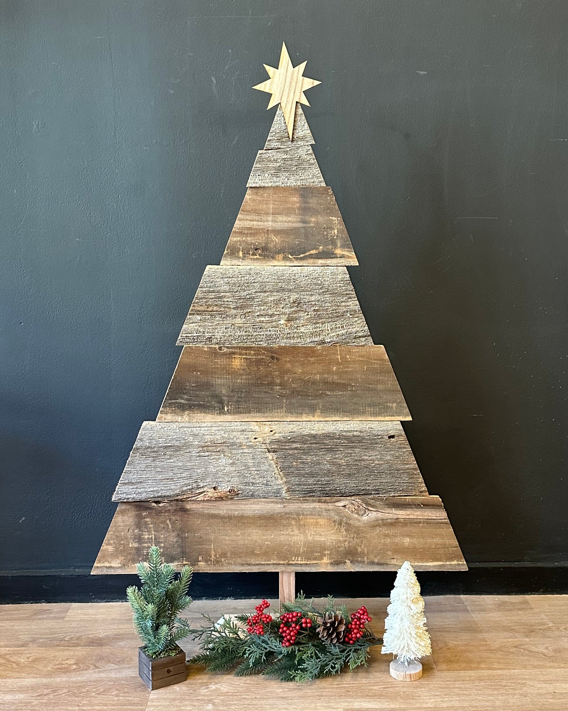 Wooden Christmas tree against a black wall with a star on top, next to a decorated Christmas tree on a wooden floor.