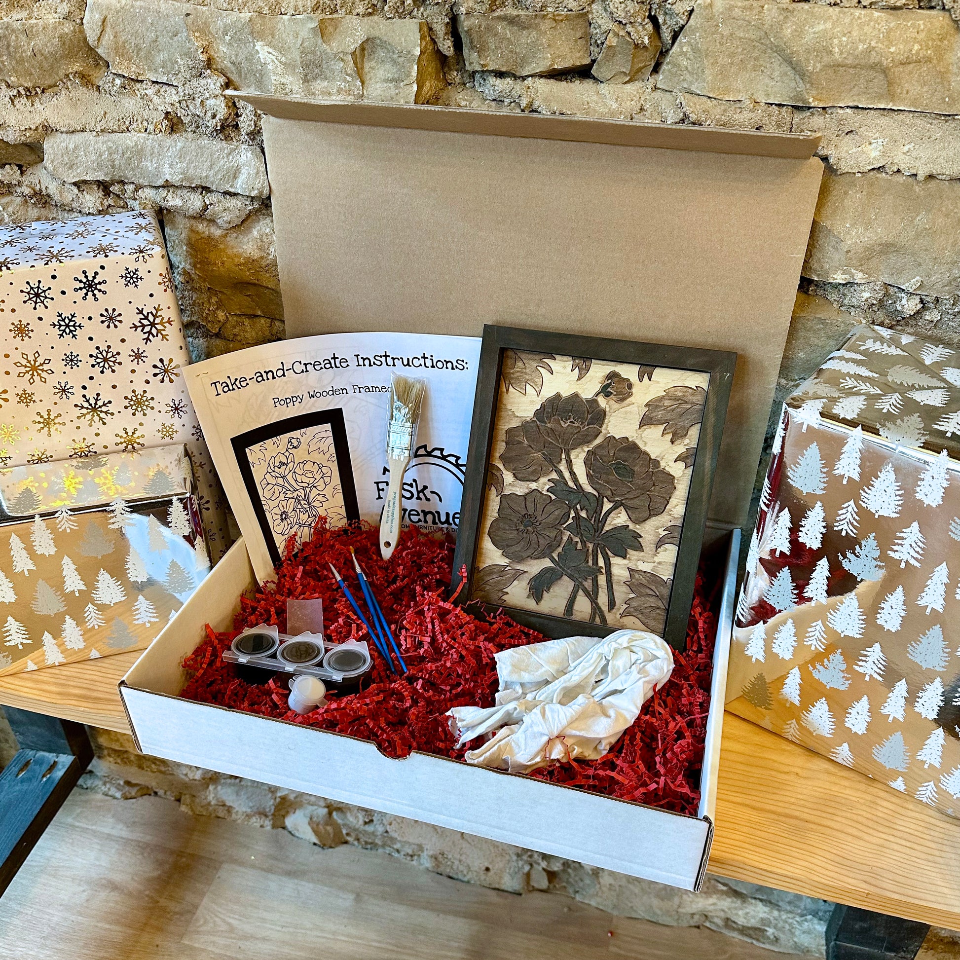 Box containing art supplies, instructions, and a wooden framed floral project against a stone wall background.