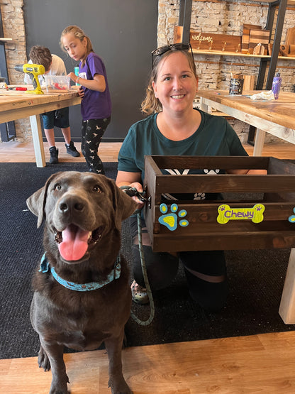 A person sitting with a brown dog in front of a wooden pet toy crate with cutouts in the shape of a paw print and bone. 2 children are standing in the background.