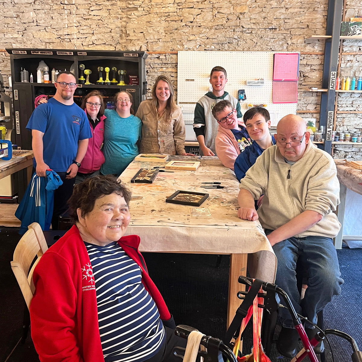 A group of people posing together in a rustic studio with stone walls, wooden tables with flower art, and shelves filled with colorful items. 