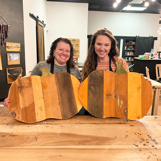 Two women holding wooden pumpkin-shaped decorations in a workshop setting.