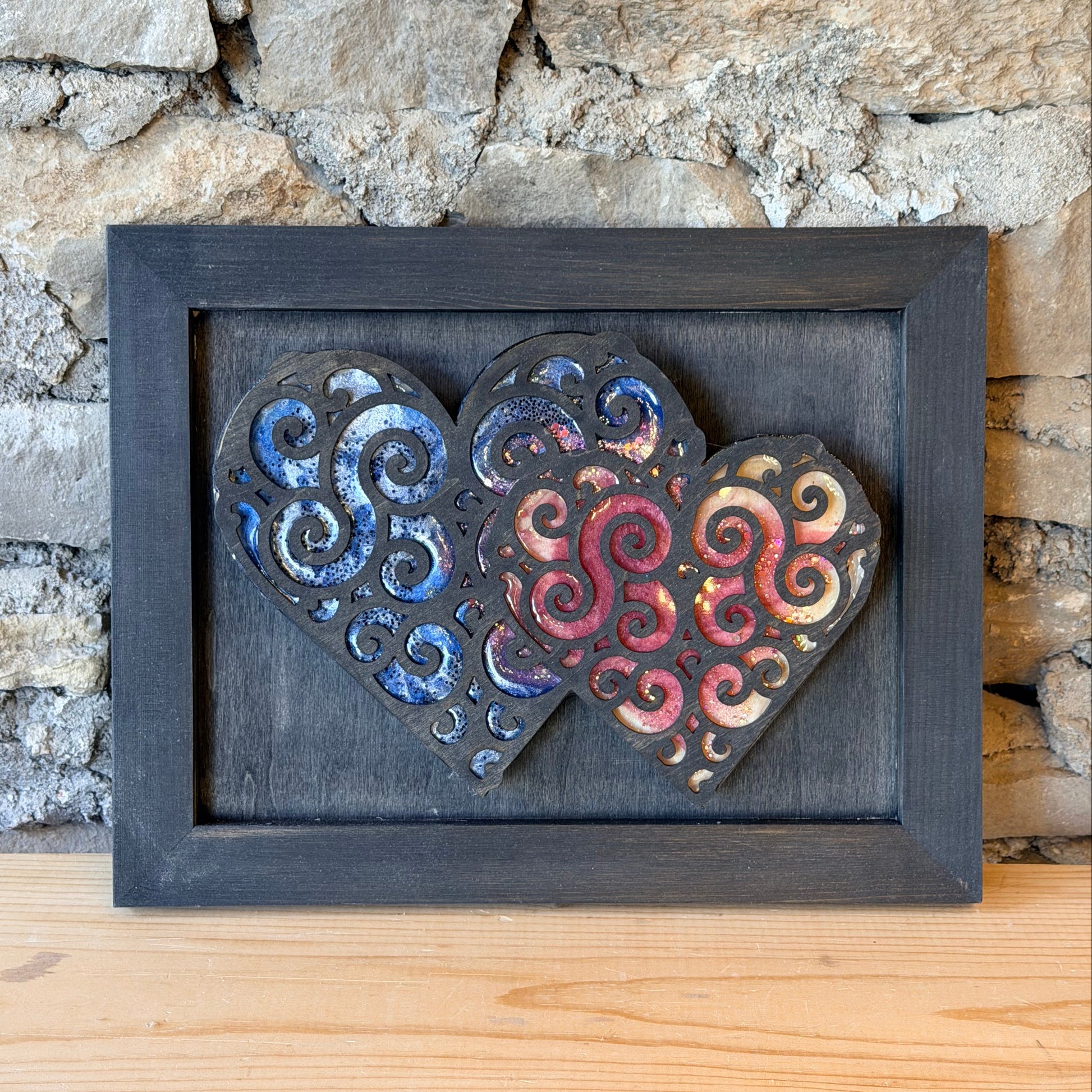 Decorative wall art with swirling patterns in hearts in a black frame against a stone wall.