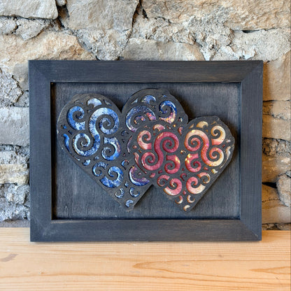 Decorative wall art with swirling patterns in hearts in a black frame against a stone wall.