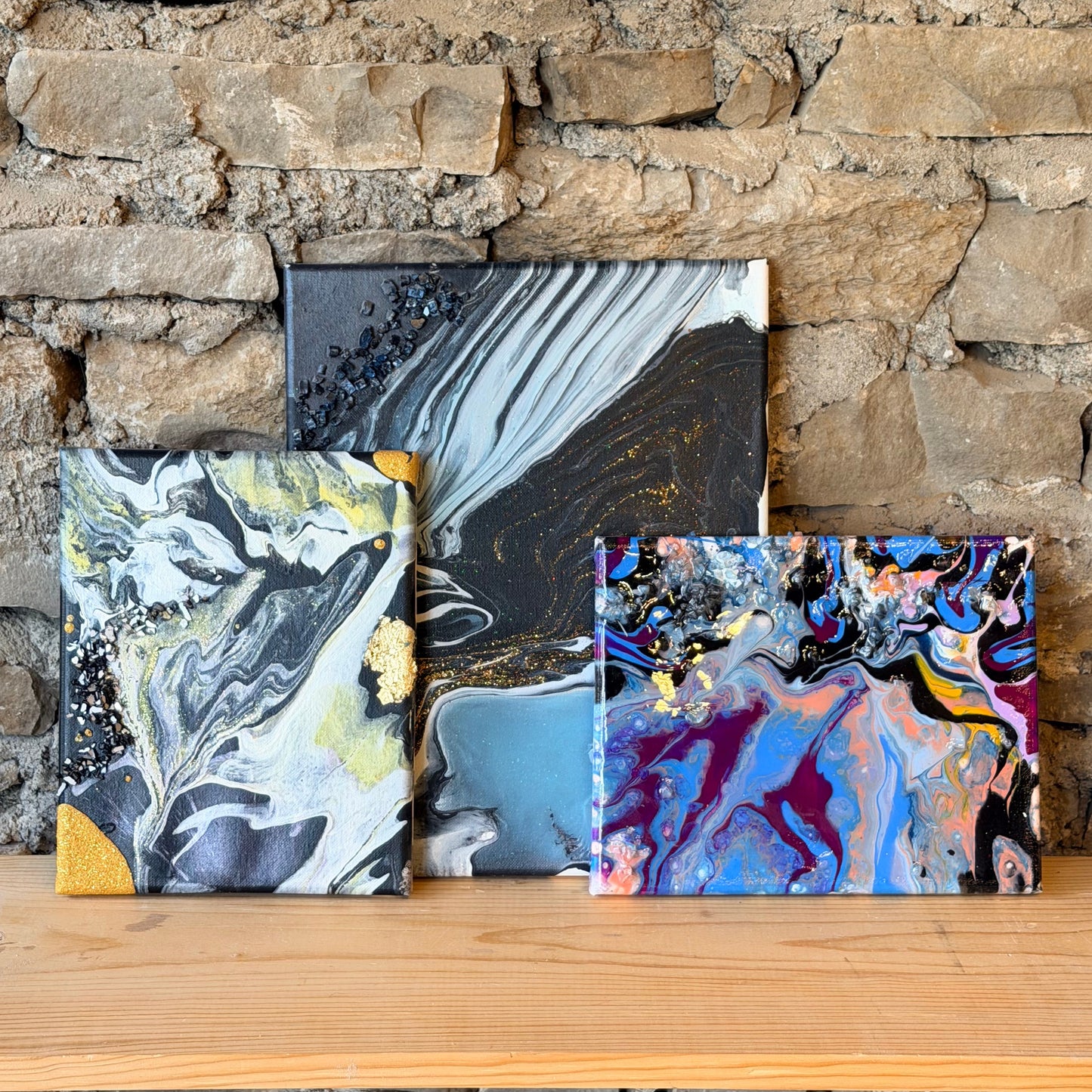 Three abstract fluid art paintings displayed against a stone wall.
