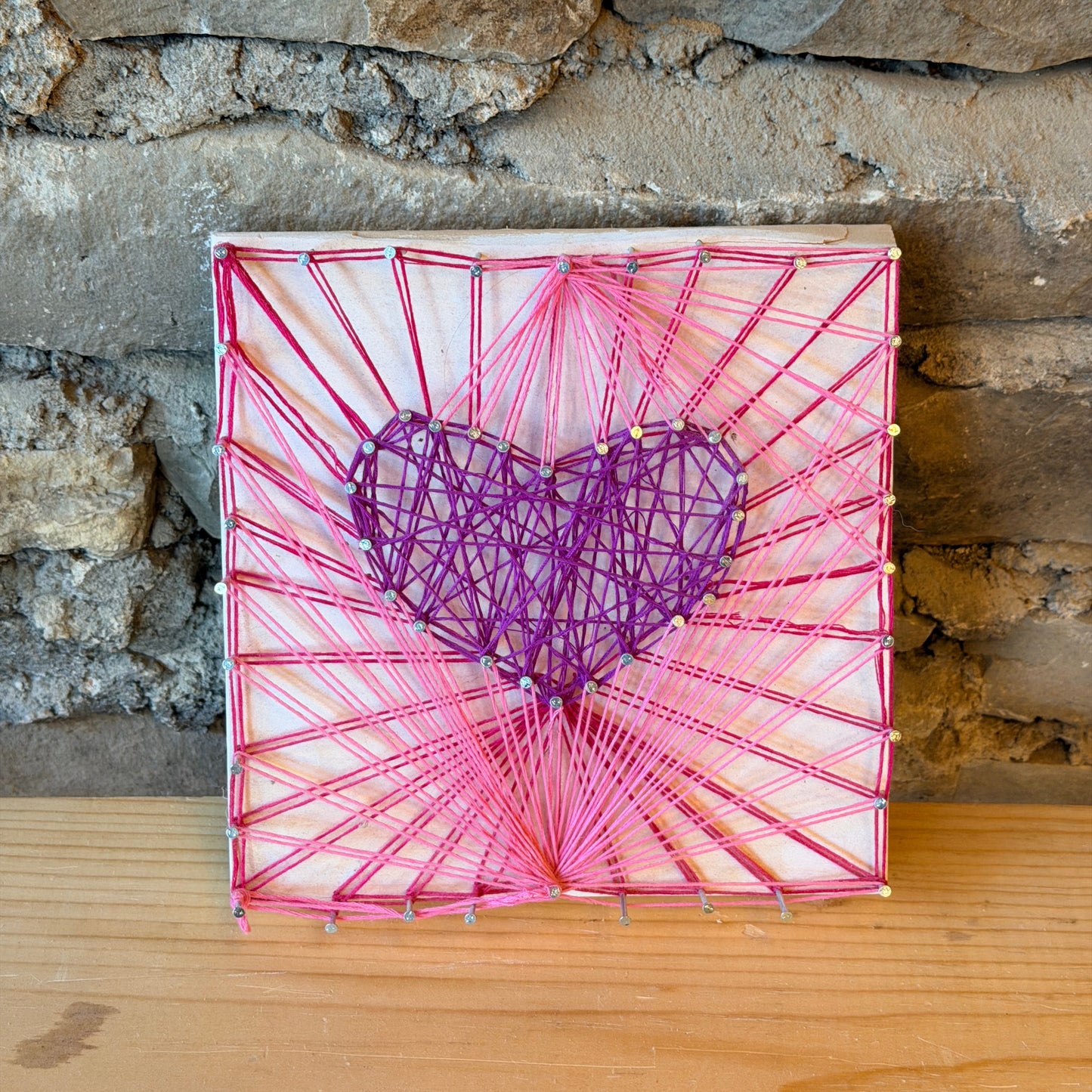 String art heart on a wooden board against a stone wall.