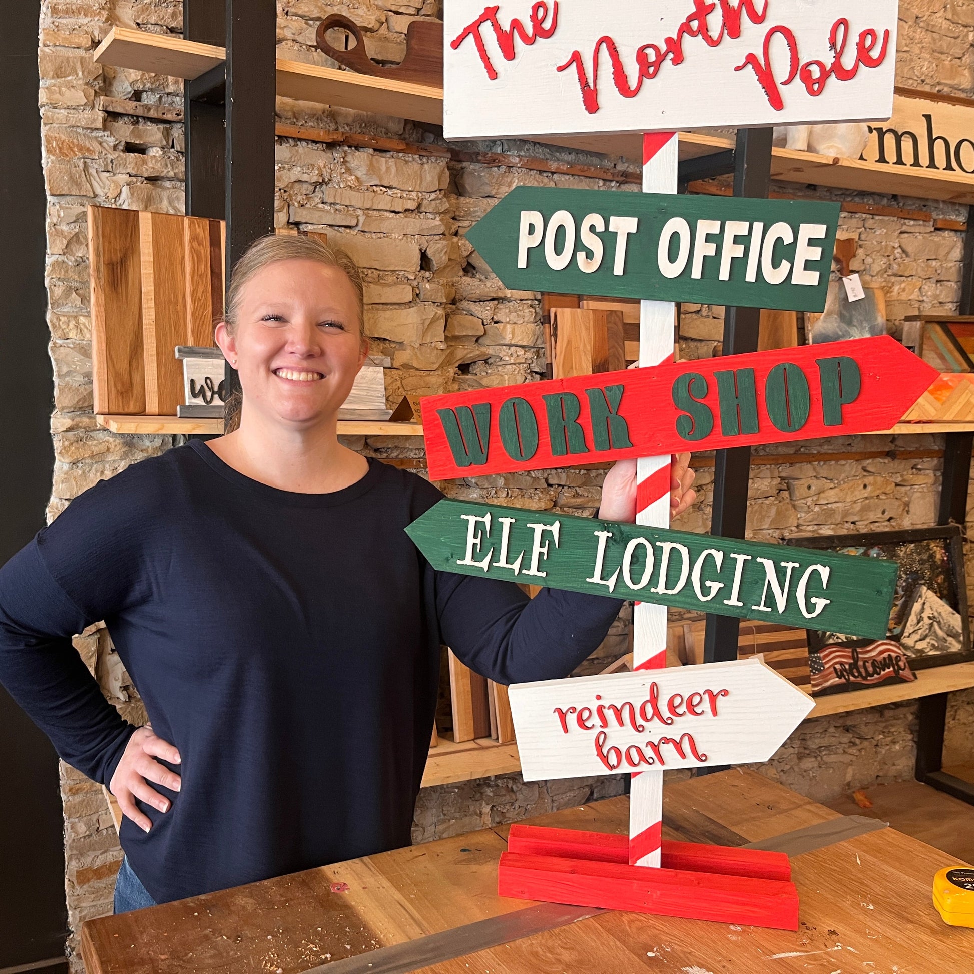 A woman standing behind a multi-directional sign that has various text labels such as 'THE NORTH POLE' and 'ELF LODGING' with red and green color scheme on a wooden surface in a workshop setting.