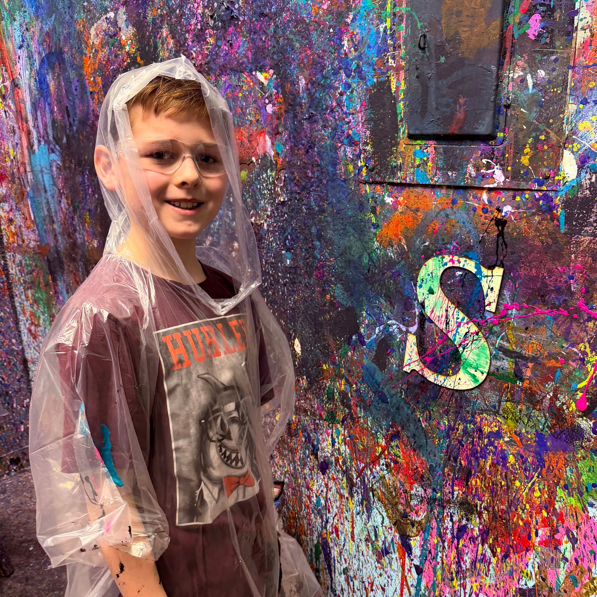 Child in a raincoat standing in front of a colorful, paint-splattered wall with a wooden letter project.