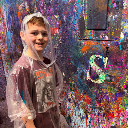 Child in a raincoat standing in front of a colorful, paint-splattered wall with a wooden letter project.