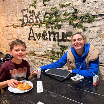 Two people sitting at a table with pizza and juice in front of a stone wall with 'Fisk Avenue' sign.