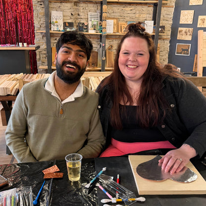 Two people sitting at a table with copper embossing art supplies and a drink, in a studio with stone walls and artwork.
