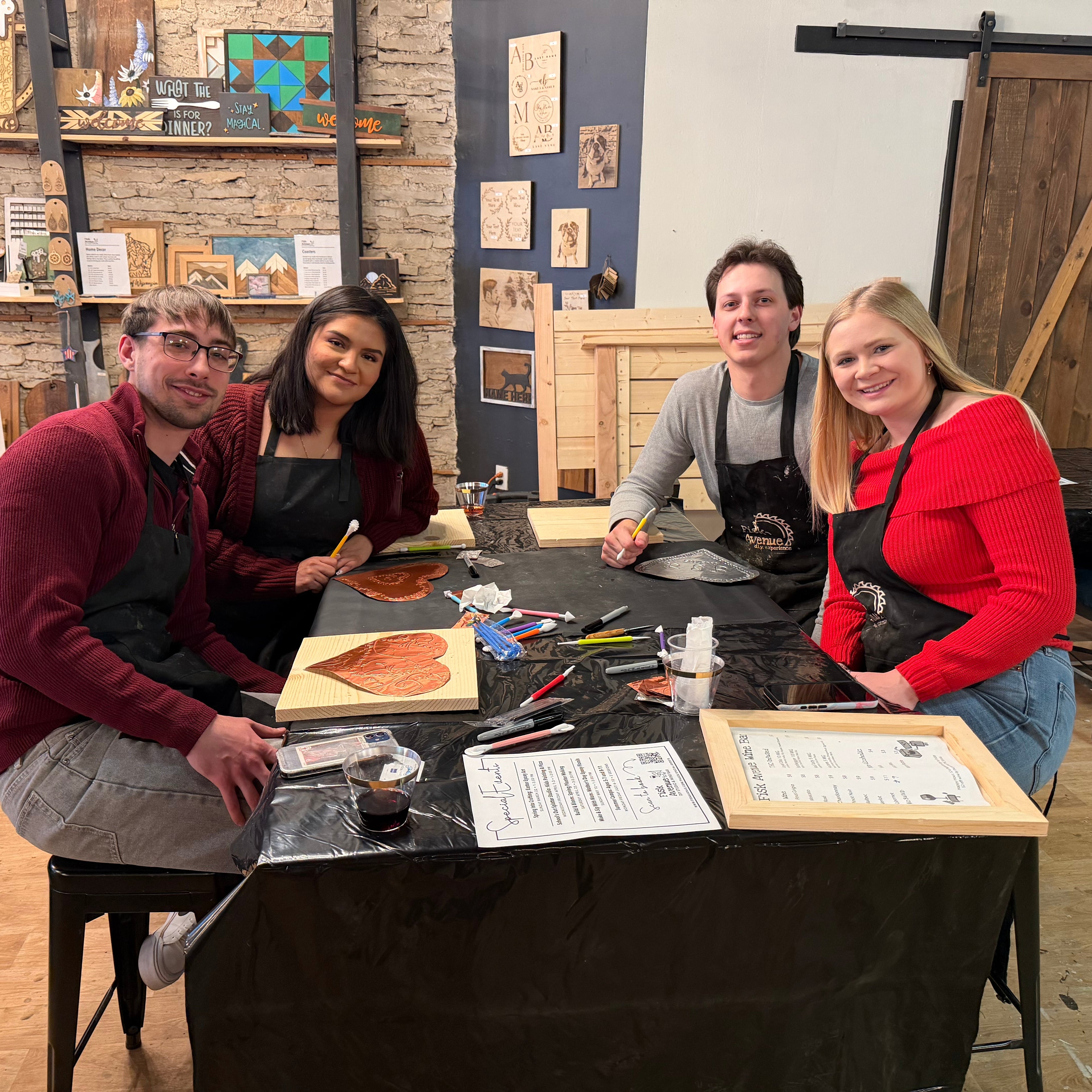 Four people in aprons standing around a table with art supplies in a workshop setting.