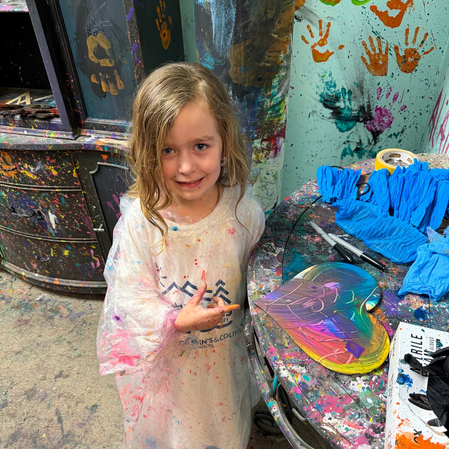 Child in a paint-splattered room with art supplies and a colorful heart painting.