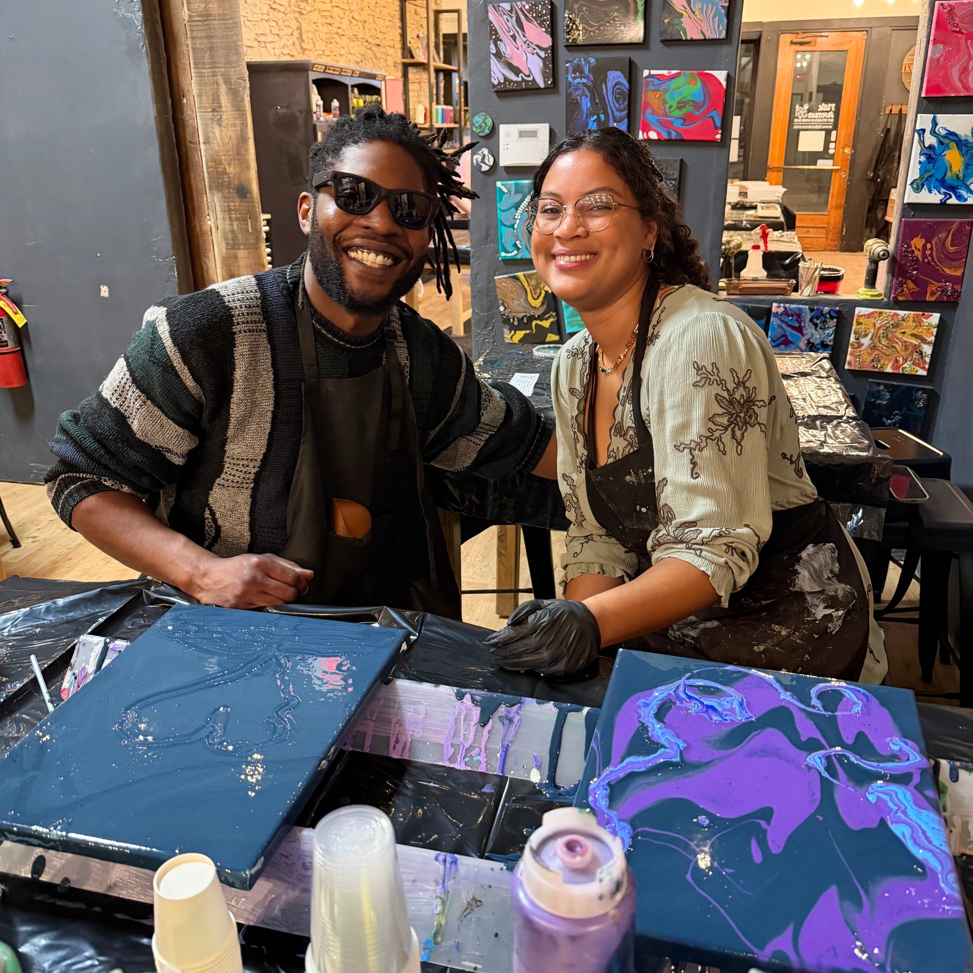 Two artists working together in a studio with art supplies and colorful paintings on the wall.