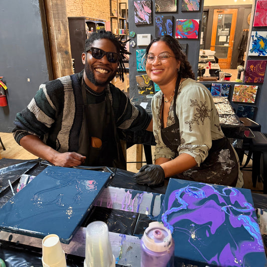 Two artists working together in a studio with art supplies and colorful paintings on the wall.