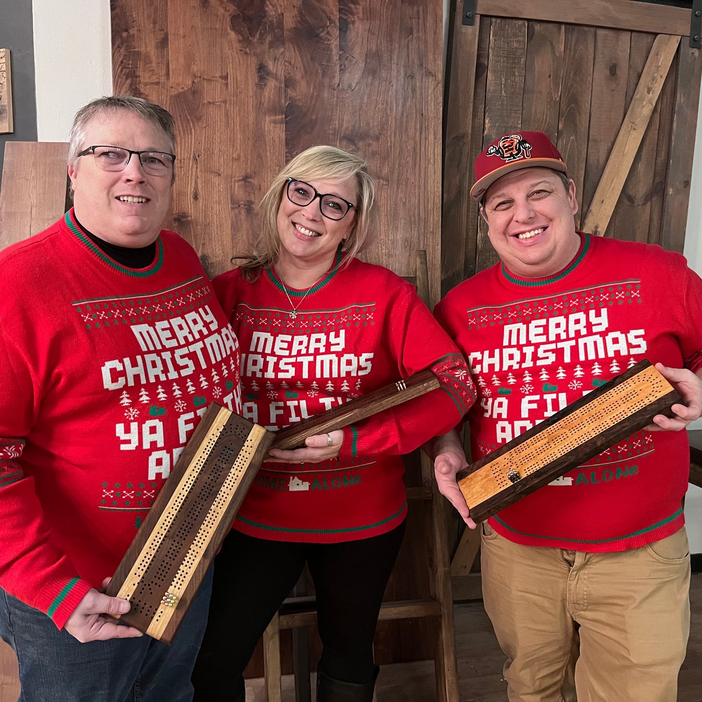 Three people wearing red 'Merry Christmas' sweaters holding wooden cribbage boards in a room with wooden walls.