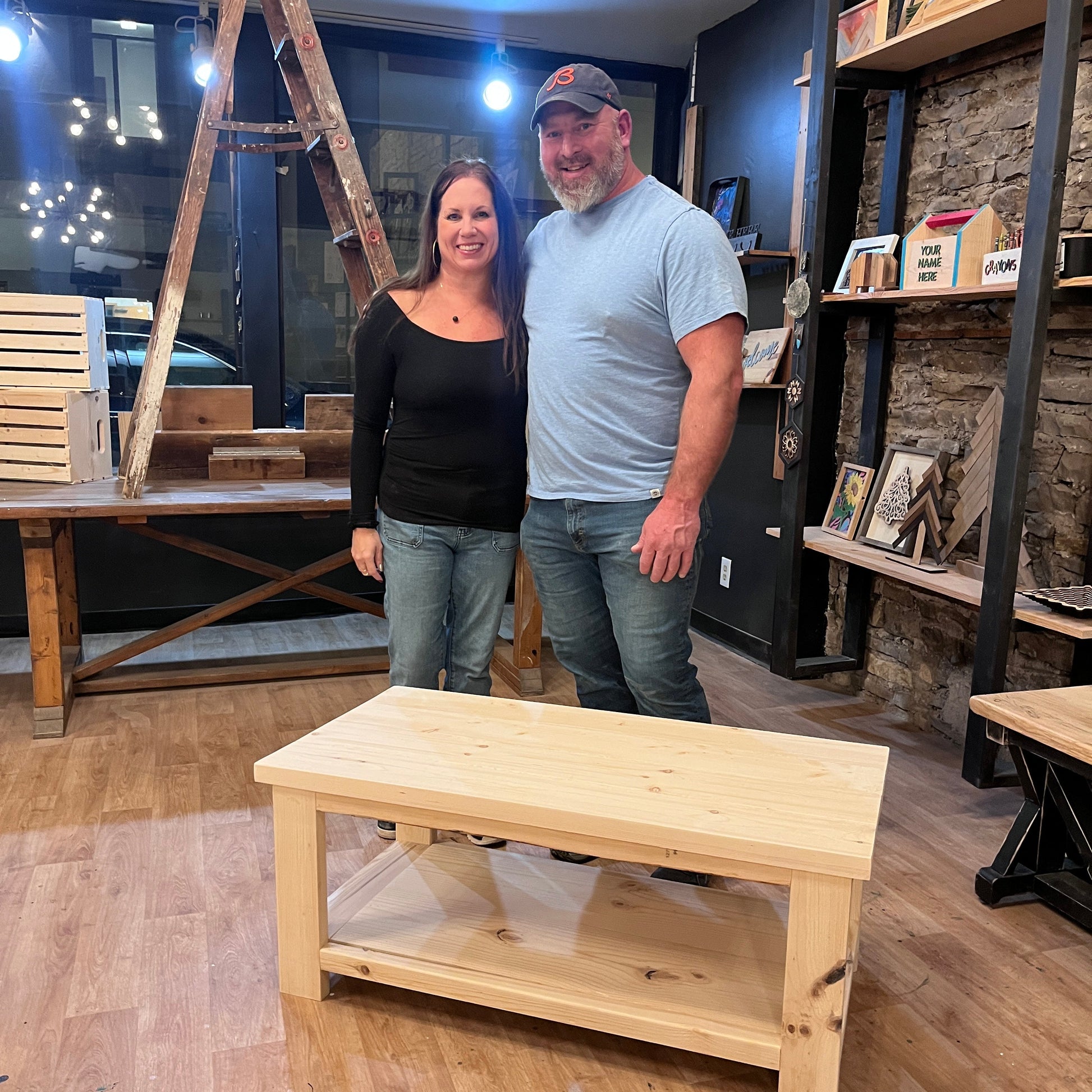 Two people standing by a wooden coffee table in a studio with wooden shelves.
