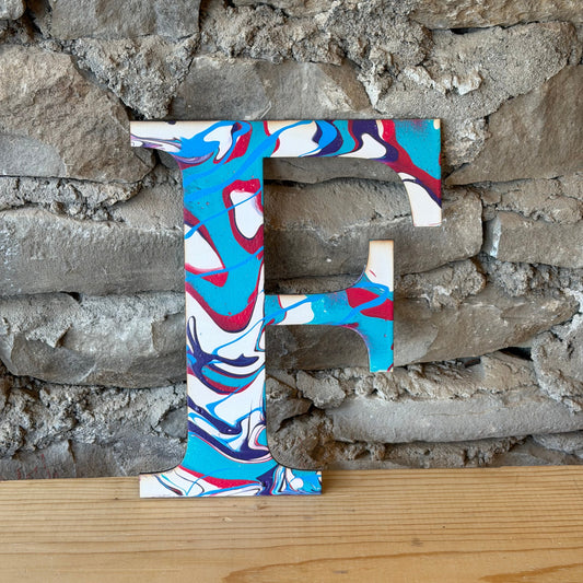 Decorative letter 'F' with a colorful fluid art pattern by a stone wall.