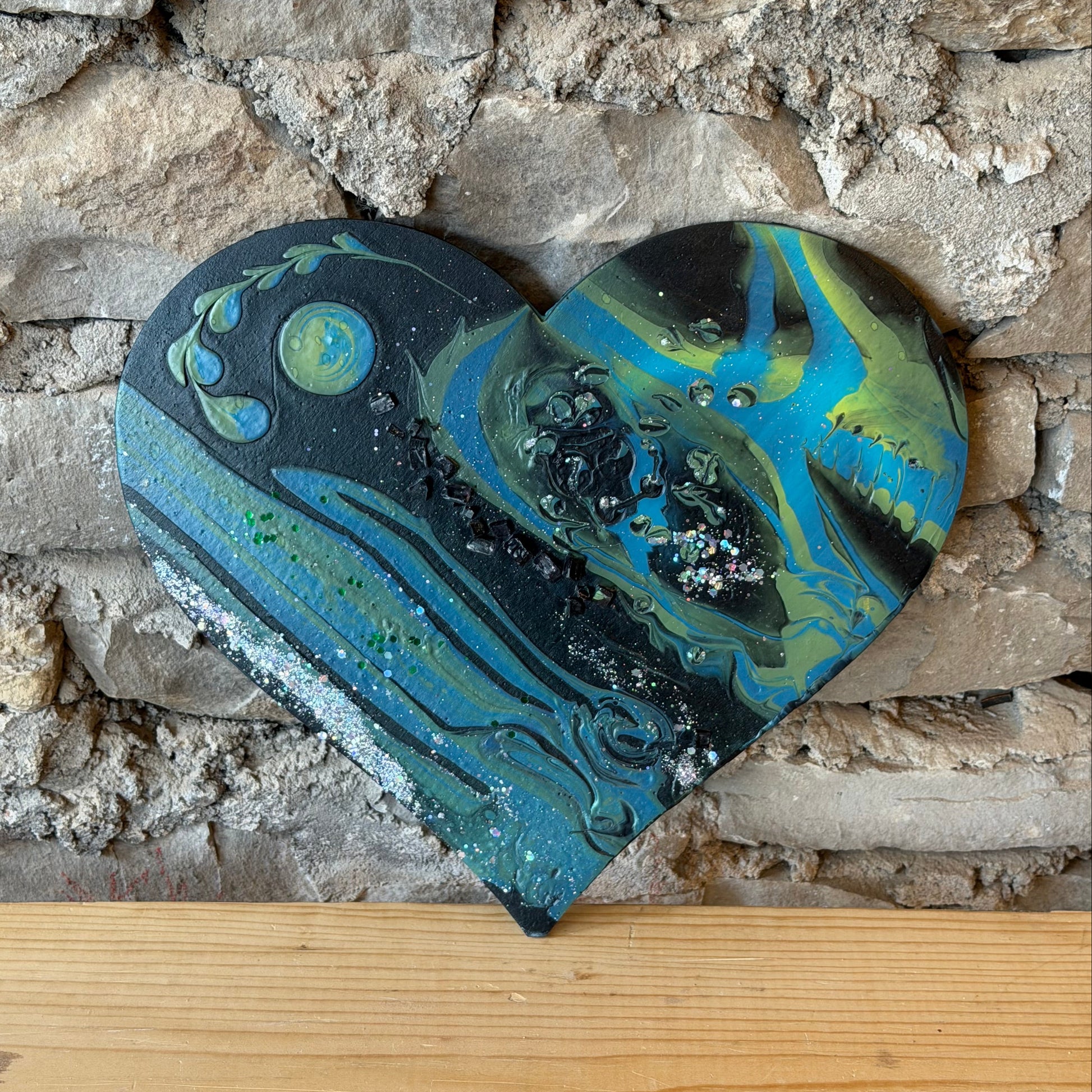 Decorative fluid art heart with blue and green patterns by a stone wall on a wooden shelf.