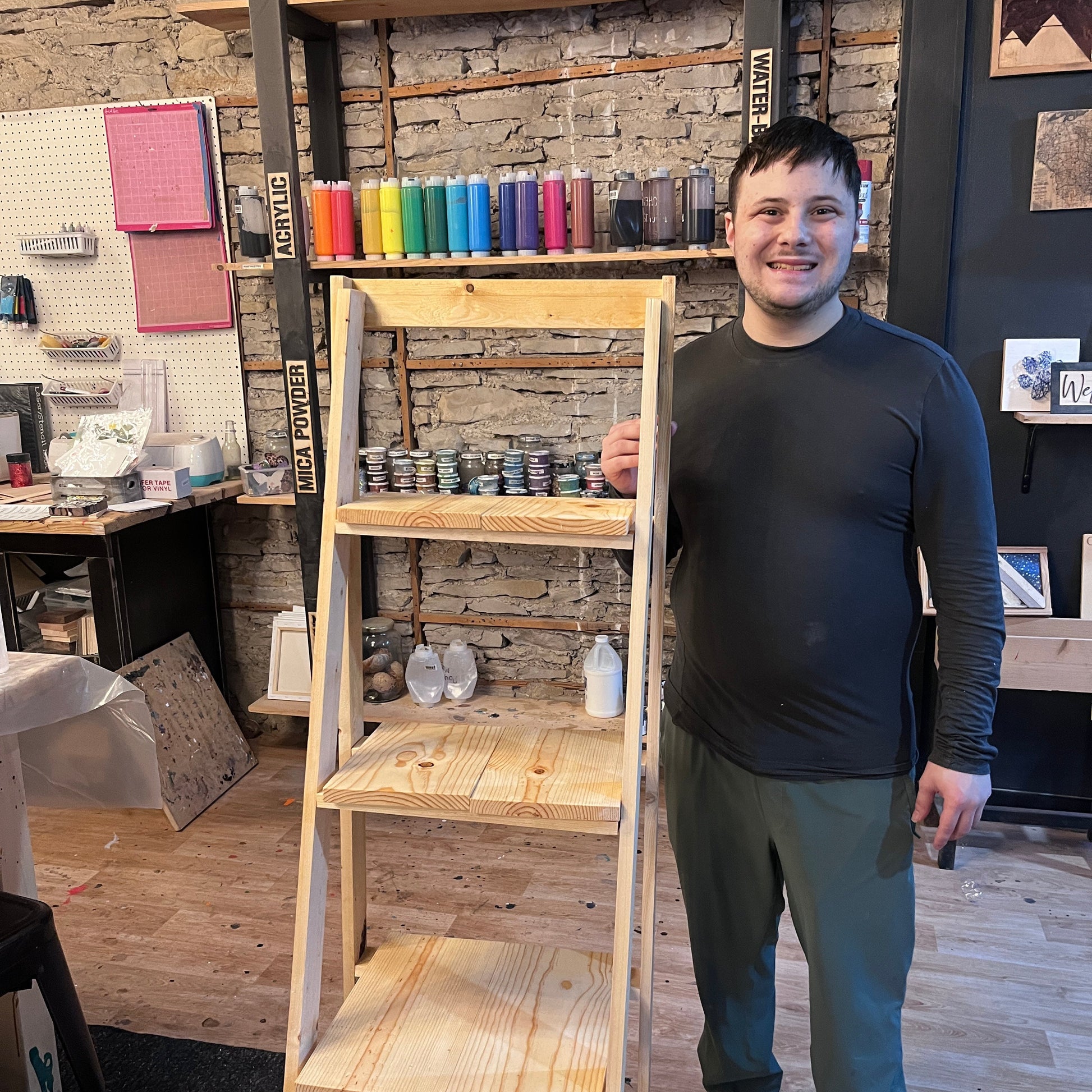 Person standing next to a wooden ladder shelf in an indoor studio setting with art supplies on shelves.