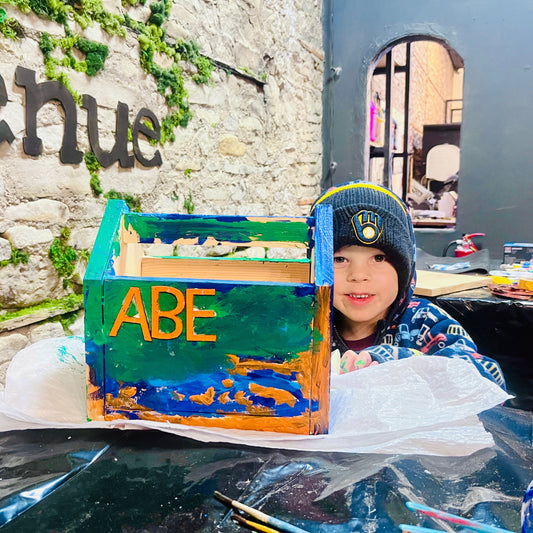 Child with a colorful wooden art caddy labeled 'ABE' in front of a stone wall.