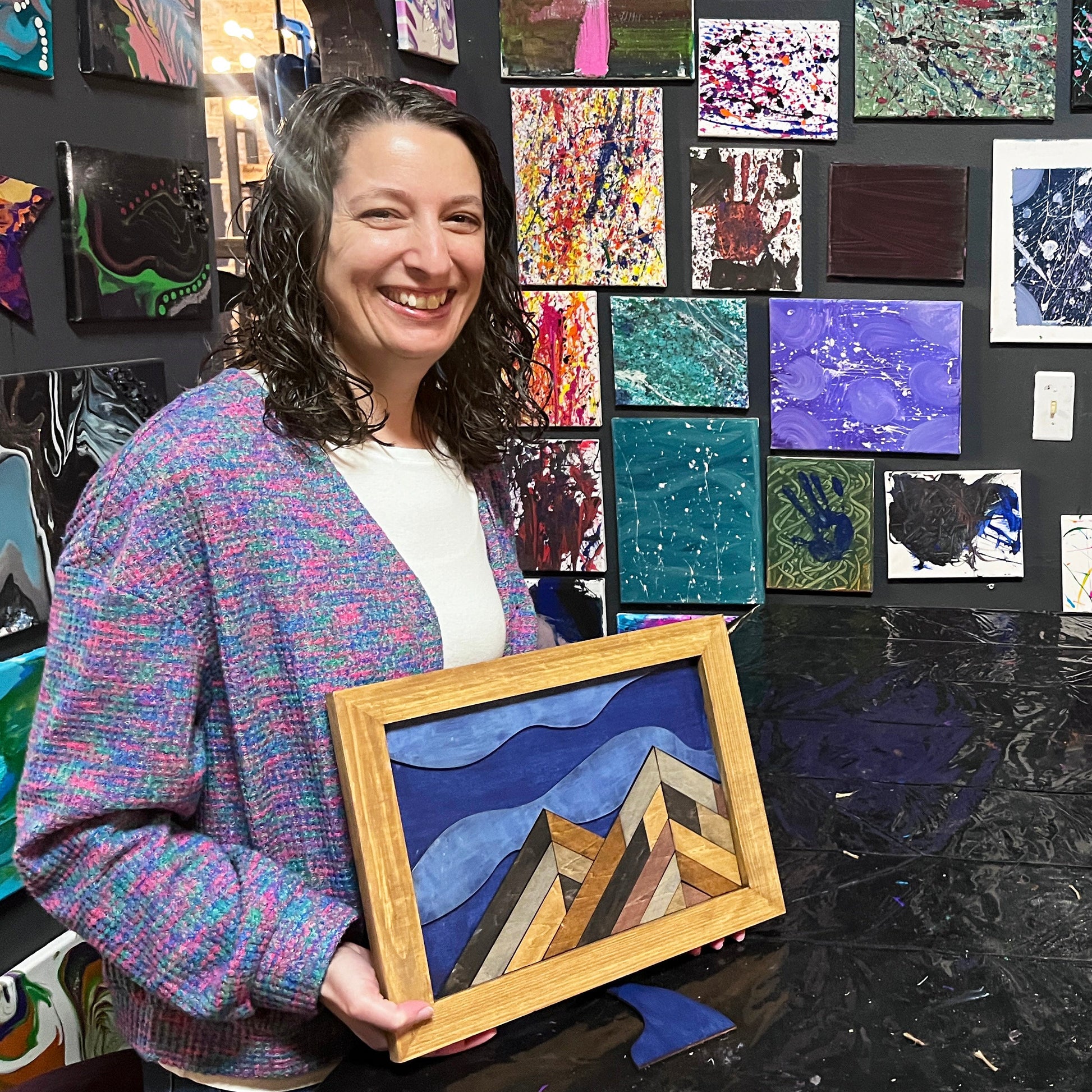 Woman standing in front of a wall of colorful art pieces, holding framed mountain artwork.