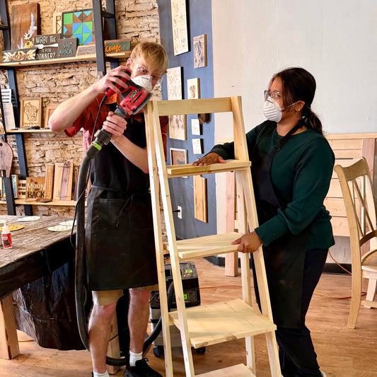 Two people working on a wooden ladder shelf in a workshop setting.
