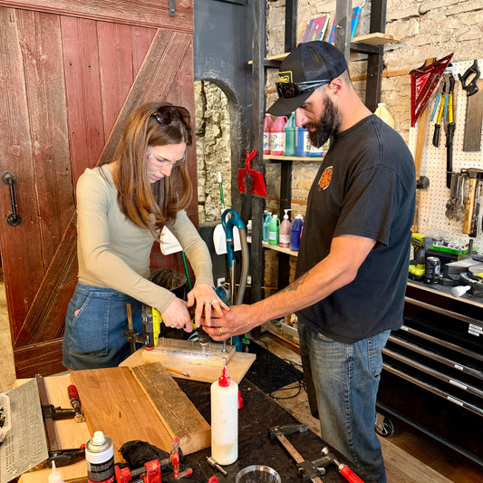 Two people working together in a cribbage board workshop with a drill and material on a table.
