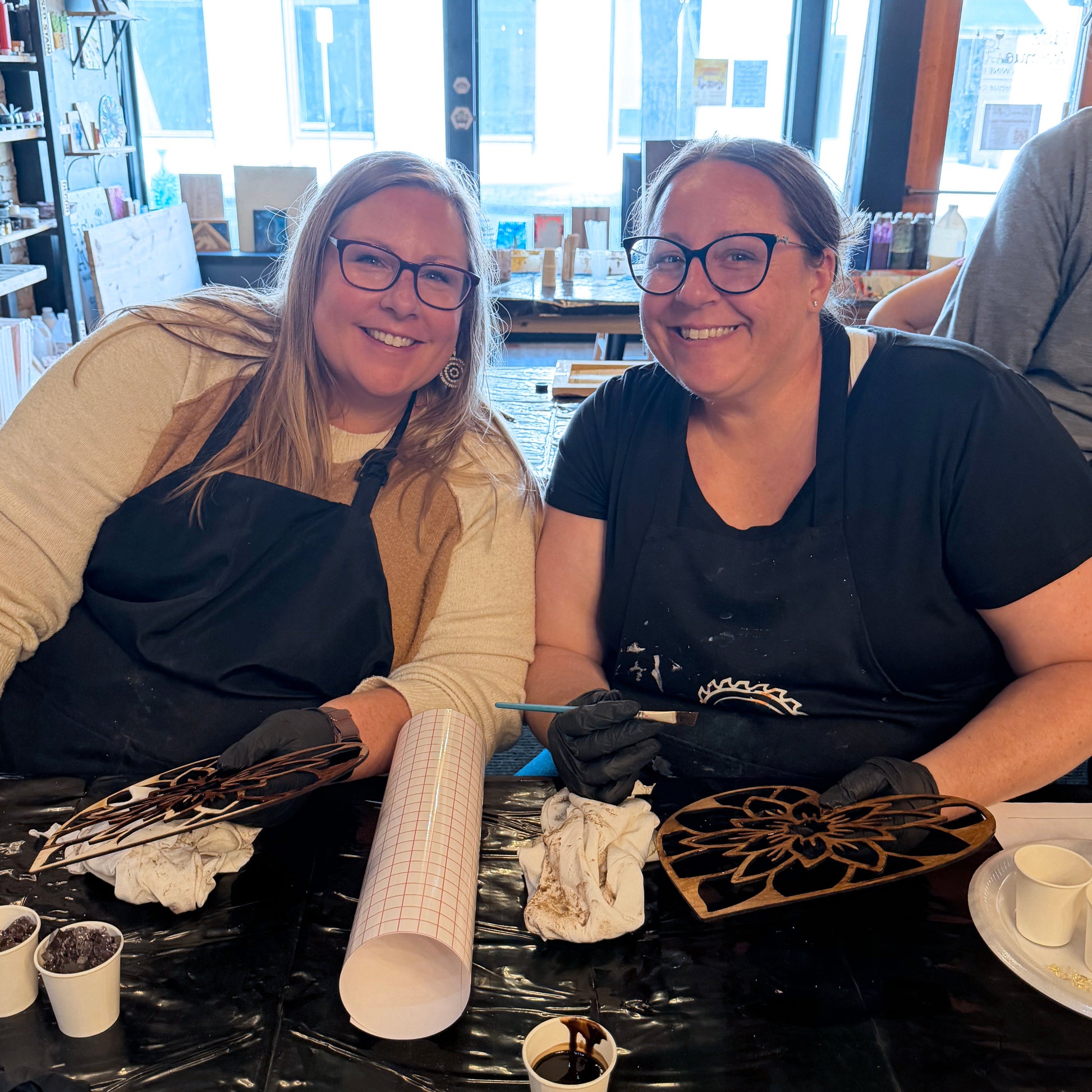 Two women sitting at a table with suncatcher art supplies, smiling.