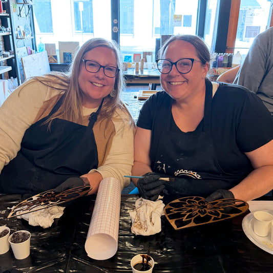 Two women sitting at a table with suncatcher art supplies, smiling.