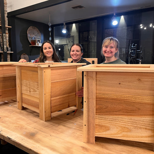 Three women standing behind wooden planter boxes in a workshop setting.