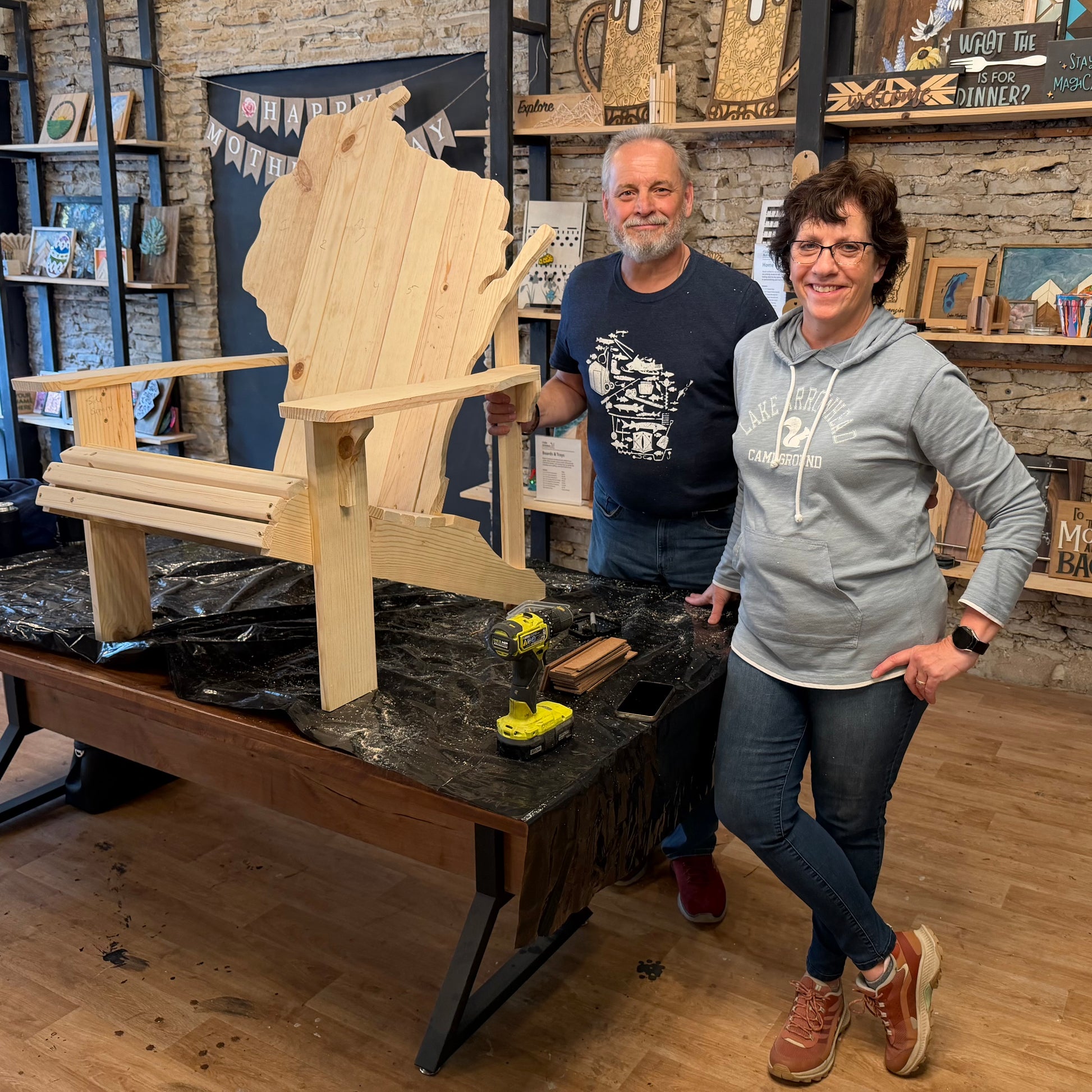 Two people standing next to a wooden chair shaped like the state of Wisconsin in a workshop.