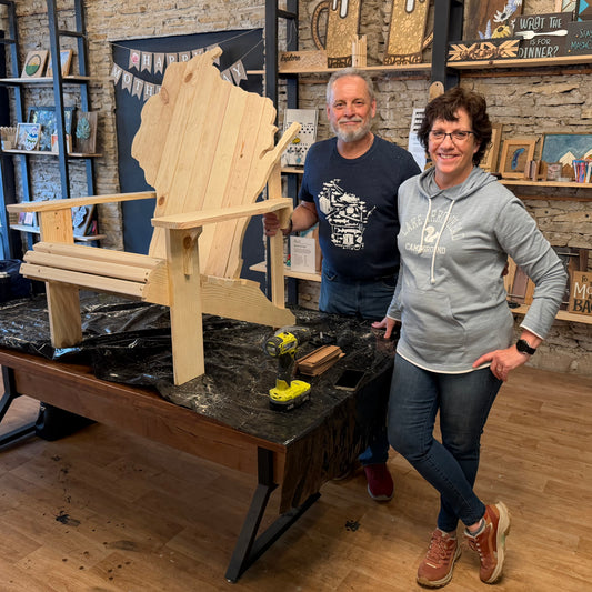 Two people standing next to a wooden chair shaped like the state of Wisconsin in a workshop.