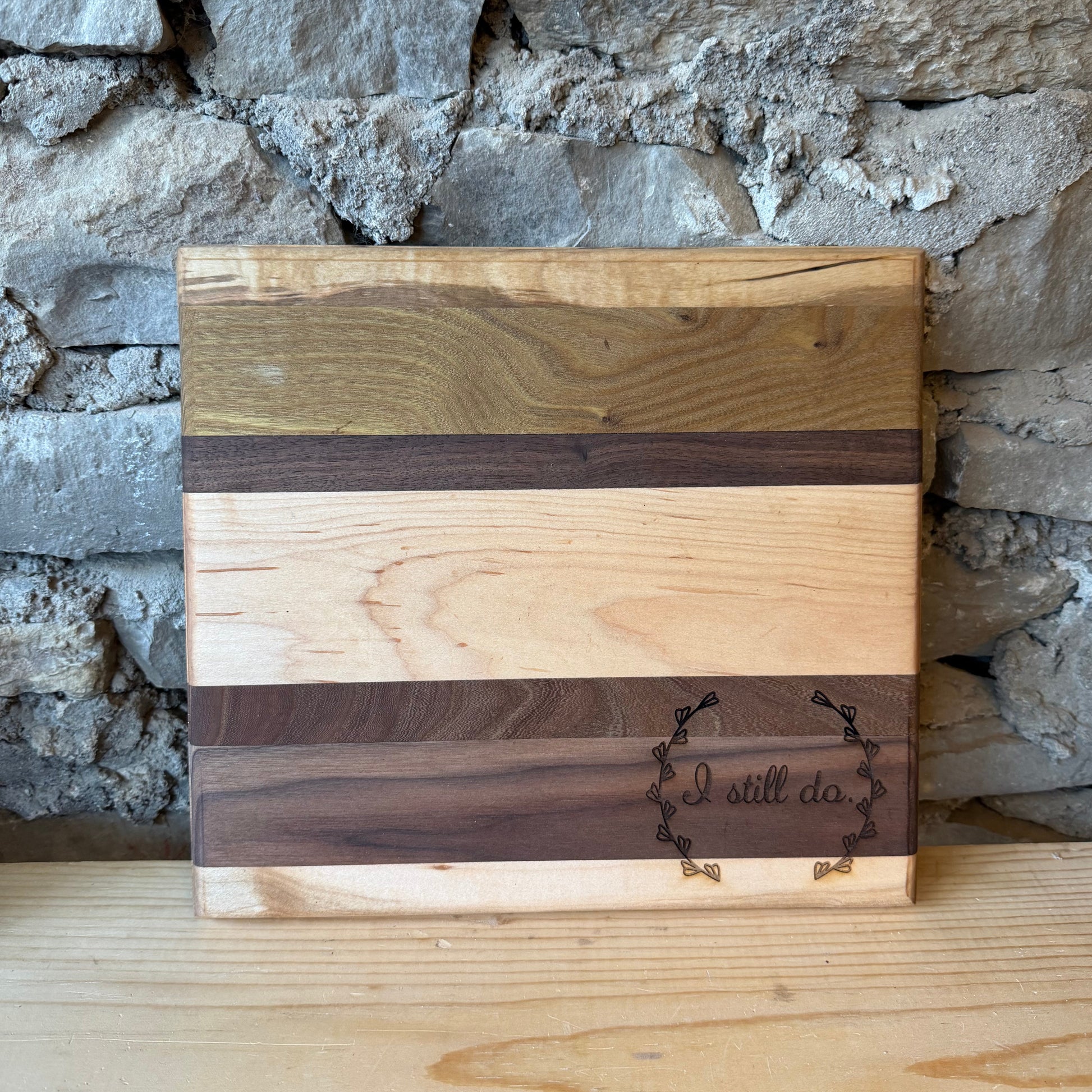 A rectangular wooden cutting board with horizontal stripes of light and dark wood grain, featuring a dark engraved logo that reads 'I still do' on a light wood surface in front of a rustic stone wall.