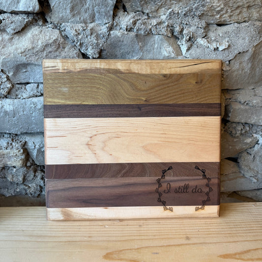 A rectangular wooden cutting board with horizontal stripes of light and dark wood grain, featuring a dark engraved logo that reads 'I still do' on a light wood surface in front of a rustic stone wall.