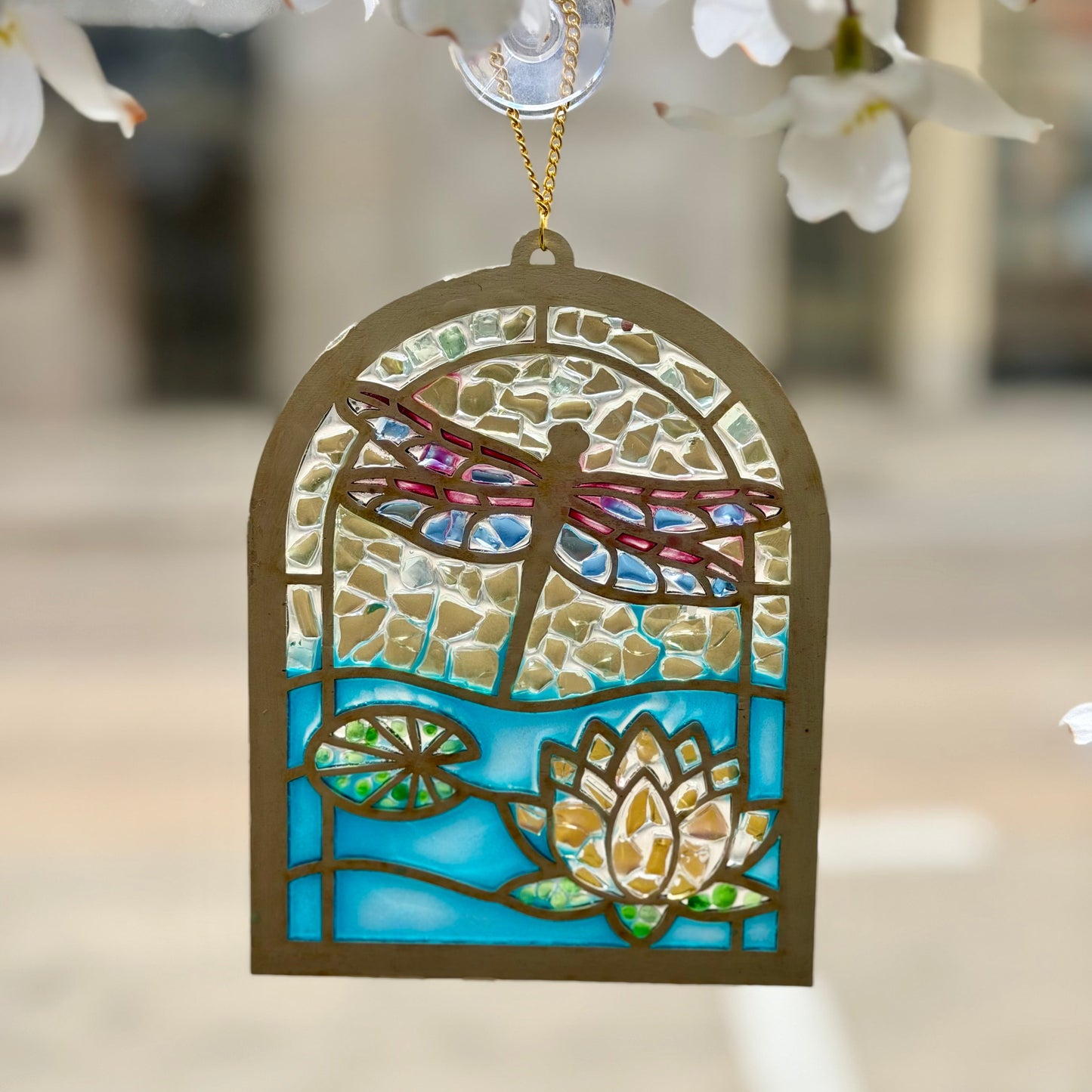 Decorative stained glass ornament with dragonfly and waterlily design, hanging against a blurred floral background.