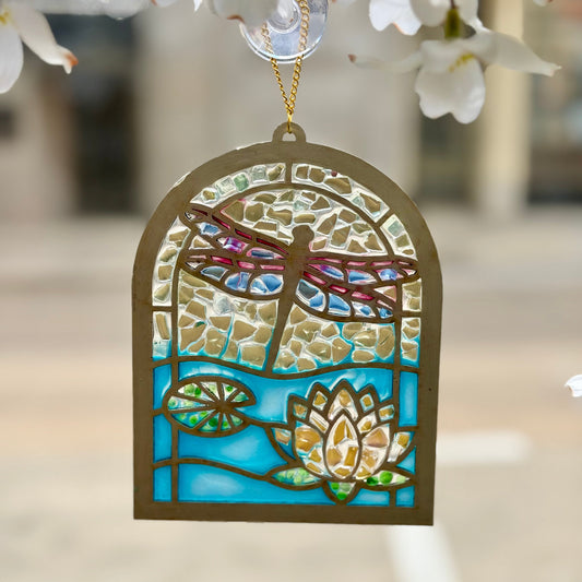 Decorative stained glass ornament with dragonfly and waterlily design, hanging against a blurred floral background.
