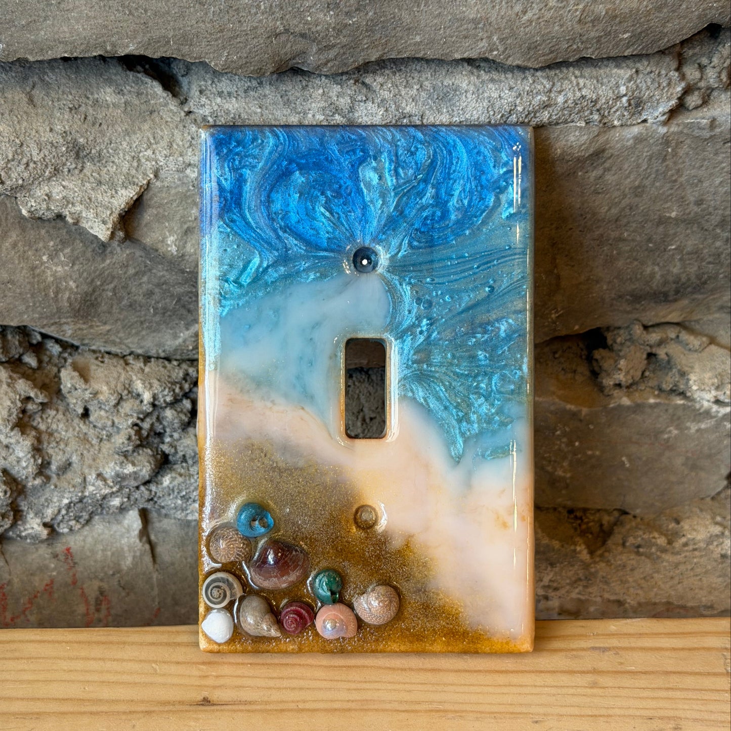 Decorative light switch cover with blue and beige beach design on a stone wall background