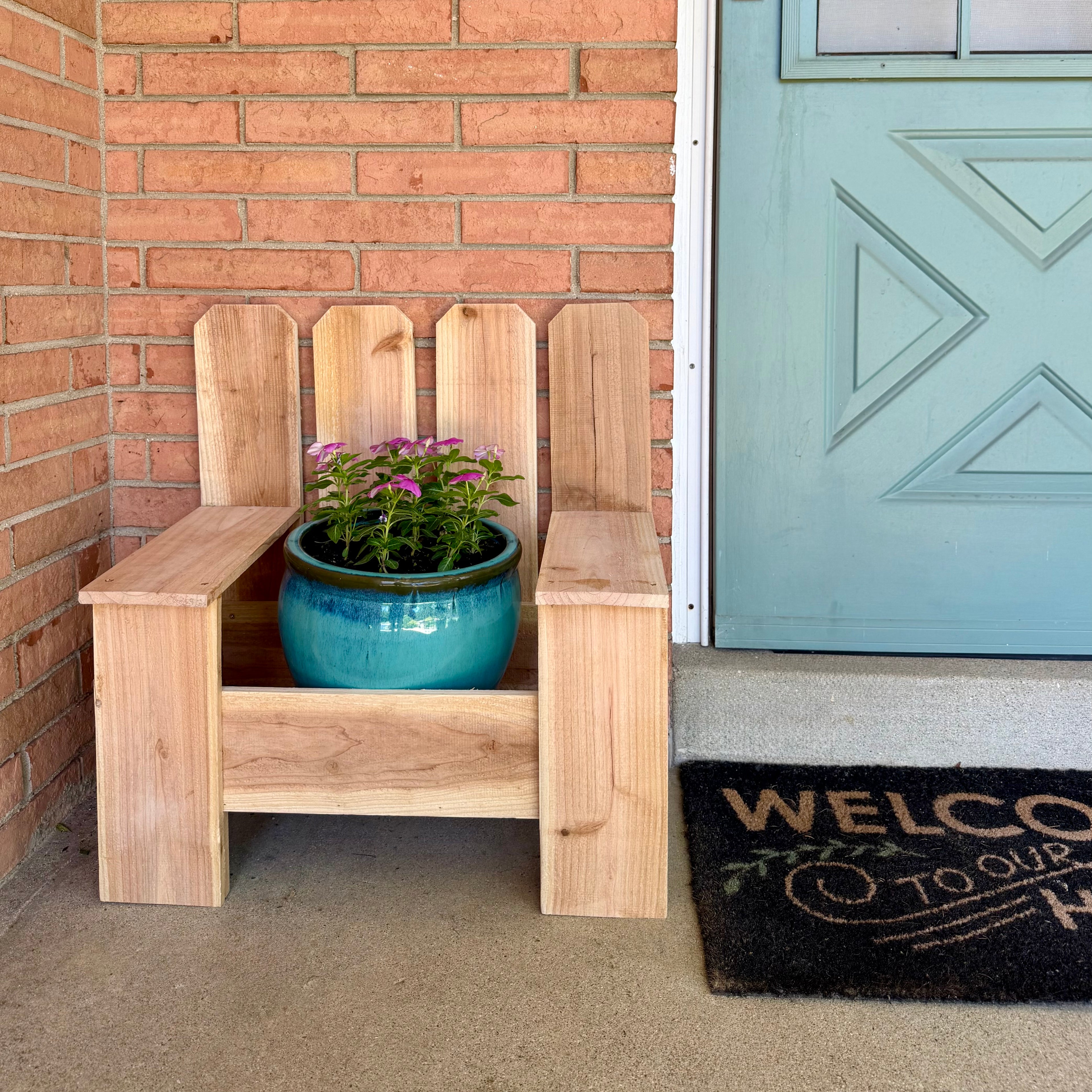 Wooden planter chair with a potted plant against a brick wall and teal door.