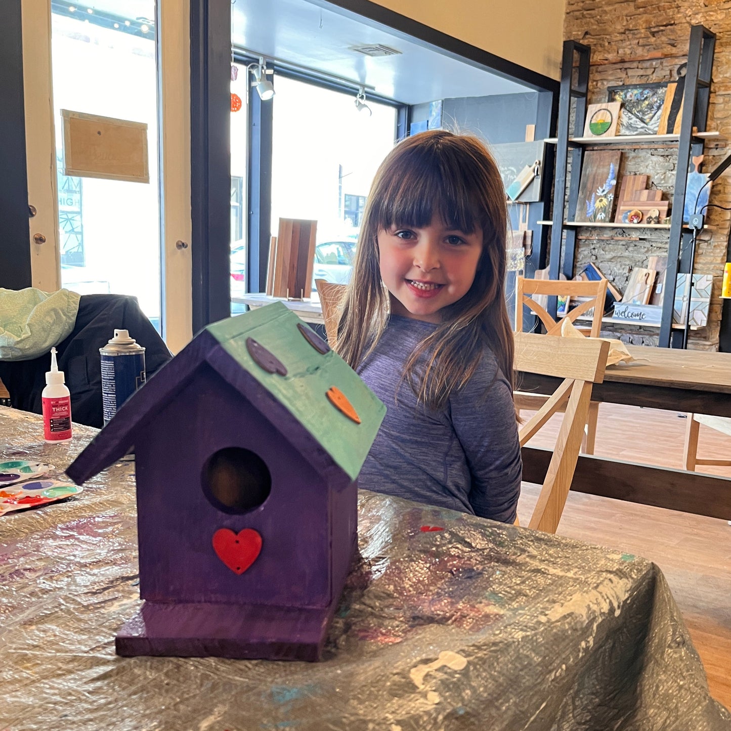 Child with a purple birdhouse decorated with a green roof and painted hearts on a table in a studio workshop.