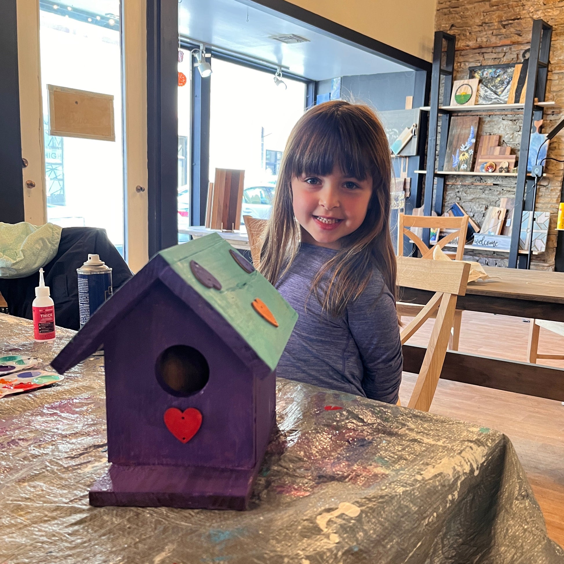 Child with a purple birdhouse decorated with a green roof and painted hearts on a table in a studio workshop.