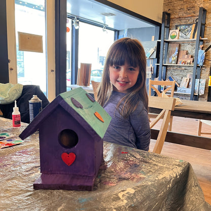 Child with a purple birdhouse decorated with a green roof and painted hearts on a table in a studio workshop.