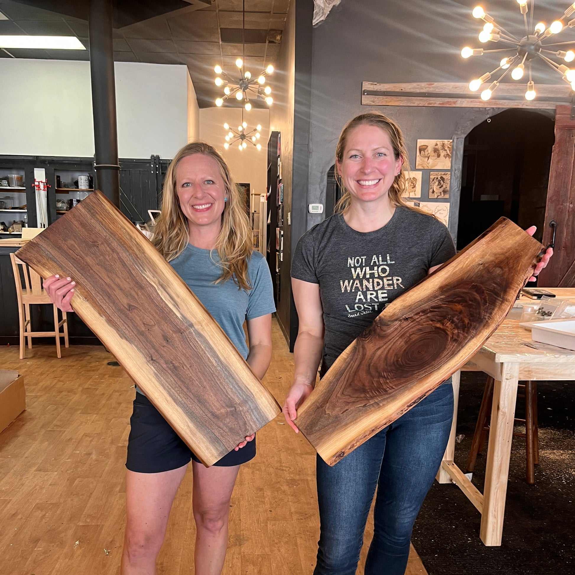 Two people holding lilve edge wooden trays in a room with wooden floors and a chandelier.