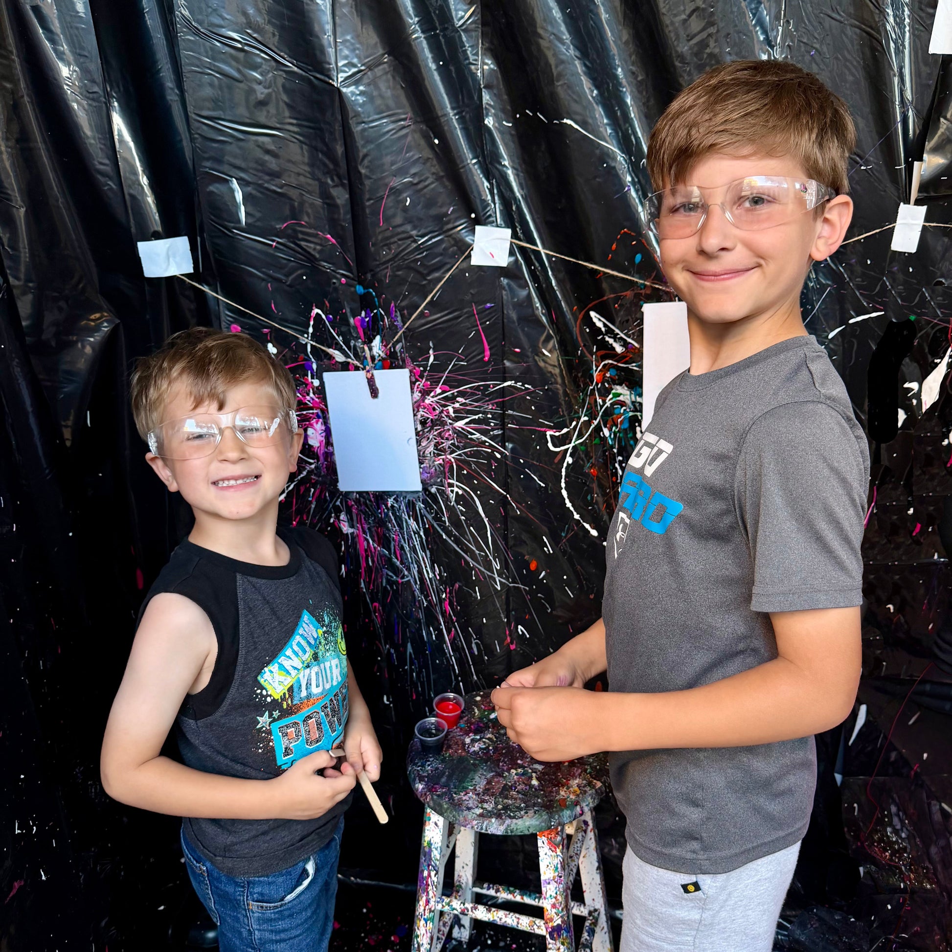 Two children with protective goggles standing in front of a black curtain with paint splatters.