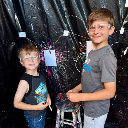 Two children with protective goggles standing in front of a black curtain with paint splatters.